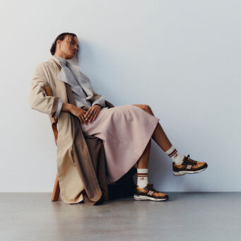 A woman sitting on a wooden chair with her legs crossed wearing a pair of New Balance Made in UK sneakers in a minimalist room with a white wall and concrete floor