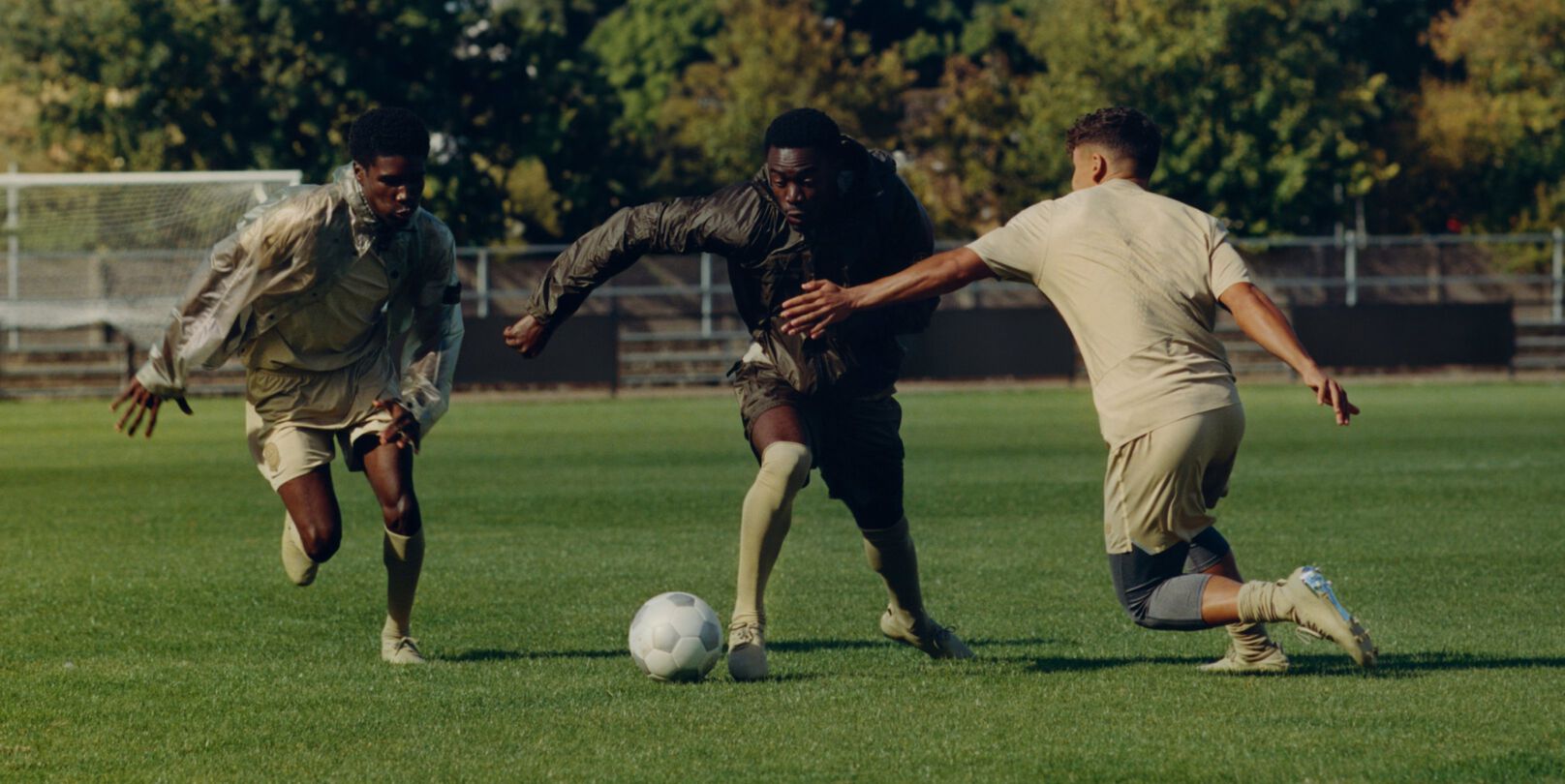 Three people playing soccer on a grassy field with a goal and trees in the background. 