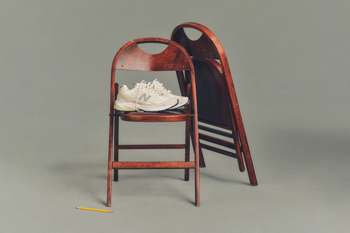 Studio image of a pair of cream-colored New Balance sneakers placed on a dark wooden folding chair, with a second matching chair folded and leaned behind it.